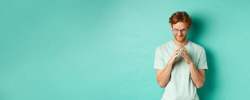 Devious redhead man in glasses and t-shirt pitching an idea, steeple fingers and look from under forehead with sly and smug smile, standing over mint background photo