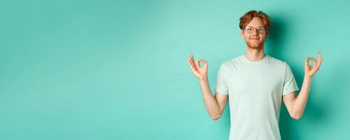 Calm and relaxed redhead man in glasses smiling, holding hands spread sideways with om mudra gesture, meditating peaceful, standing over mint background photo
