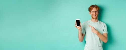 Attractive young man with red beard and hair pointing finger at blank smartphone screen, showing online promotion or app, smiling at camera, turquoise background photo