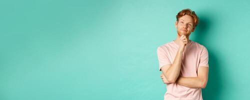 Pensive handsome man with red hair and beard looking at upper left corner, making choice and looking thoughtful, standing in t-shirt over mint background photo