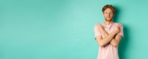 Attractive indecisive man in t-shirt cross hands and pointing sideways, showing two choices, need help with decision, standing over turquoise background photo