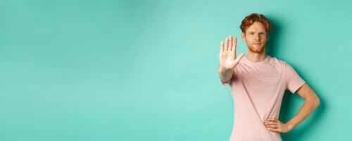 Serious young man with red hair and beard telling to stop, showing palm and frowning, disapprove and prohibit something bad, standing over turquoise background photo