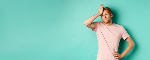 Annoyed and bothered redhead male model showing facepalm and roll eyes, standing over mint background photo