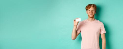 Handsome young man smiling and showing plastic credit card, standing in t-shirt against turquoise background photo