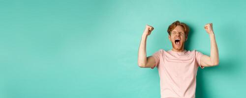 Young redhead man triumphing as watching sport game, making bet on winning team and rejoicing, screaming with joy and satisfaction, standing over turquoise background photo