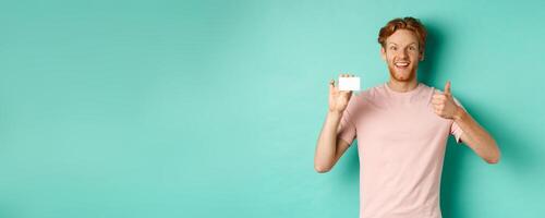 Cheerful male bank client in t-shirt showing thumb up and plastic credit card, smiling satisfied at camera, standing over turquoise background photo