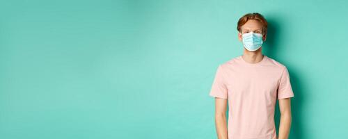 Covid-19, social distancing and quarantine concept. Young guy with red hair wearing medical mask from coronavirus in public, standing over turquoise background photo