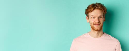 Close up of redhead bearded man looking pleased, nod in approval and smiling, standing in pink t-shirt against turquoise background photo