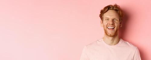 Close up of cheerful young man with messy red hair and beard, laughing and smiling, showing white teeth, looking happy at camera, standing over pink background photo