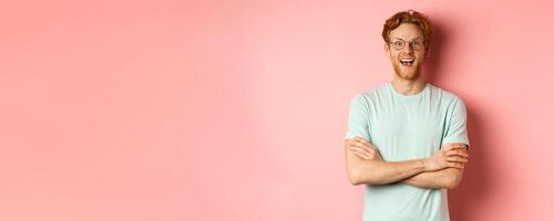 Portrait of cheerful european man in glasses looking amazed at camera, see interesting promotion, standing amused over pink background photo