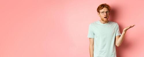 Portrait of handsome redhead man in glasses looking confused, open mouth and stare at camera, cant understand nothing, standing over pink background photo