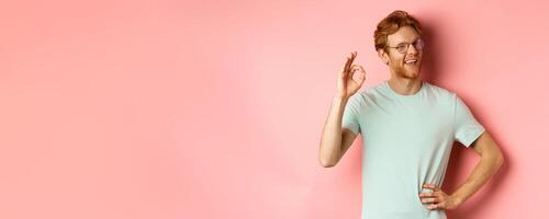 Cheerful guy with red hair and beard, wearing glasses, showing OK sign in approval and winking, smiling satisfied, standing over pink background photo