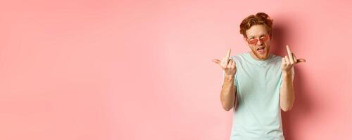 Arrogant and careless redhead man in sunglasses dont give a fuck, showing middle fingers at camera and frowning, standing over pink background photo