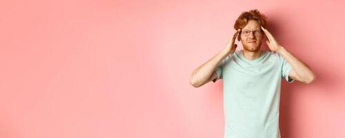 Portrait of redhead man in crooked glasses touching head and feeling dizzy or nauseous, having hangover or headache, standing over pink background photo