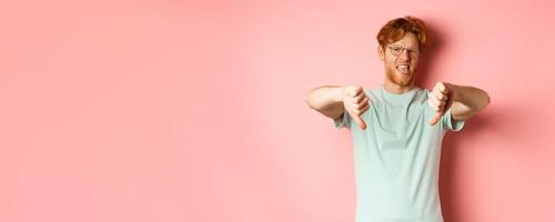 Disgusted redhead man in t-shirt and glasses showing thumbs down and tongue, grimacing from aversion, standing over pink background photo