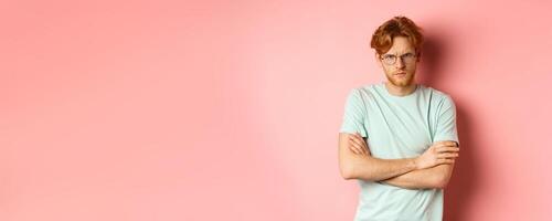 Angry man in glasses with red hair frowning, cross arms on chest in defensive pose, sulking at you, standing over pink background photo