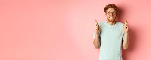 Worried redhead man waiting for results, expecting something with fingers crossed, biting finger and looking at something risky, standing over pink background photo