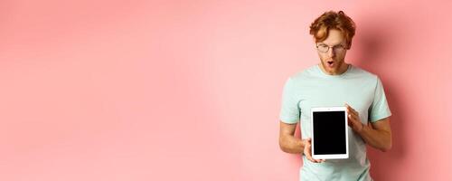 Impressed redhead man in glasses, showing blank digital tablet screen and looking in awe at display, saying wow, standing in t-shirt against pink background photo