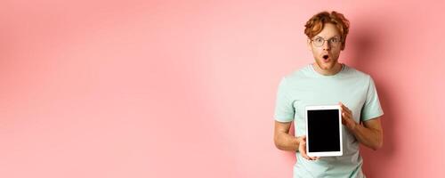Excited young man with red hair and beard, checking out online promotion, showing digital tablet screen and staring at camera amazed, standing over pink background photo