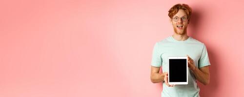 Excited young man with red hair and beard, checking out online promotion, showing digital tablet screen and staring at camera amazed, standing over pink background photo