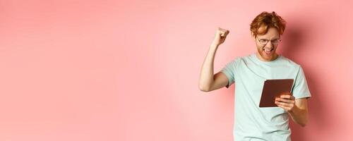 Image of happy redhead man triumphing, winning online with digital tablet and rejoicing, standing over pink background photo