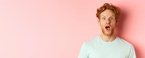 Face of surprised redhead man reacting to cool promo offer, raising eyebrows and gasping, staring in awe at camera, standing over pink background photo