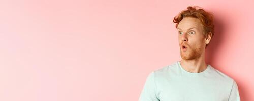 Headshot portrait of surprised redhead man with beard, looking left and saying wow, raising eyebrows amazed, standing over pink background photo