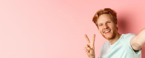 Close-up of happy redhead man taking selfie on smartphone and showing peace sign, view from phone camera, pink background photo