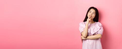 Pensive smiling asian girl making choice, looking at upper left corner logo and thinking, having an idea, standing cunning on pink background photo