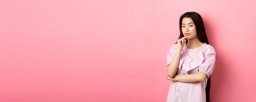 Hmm, interesting. Pensive and cunning asian girl having idea, plan to do something, looking at camera and touching chin with thoughtful face, standing on pink background photo