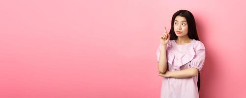Beautiful asian girl raising finger and looking left at logo, having an idea, found solution, standing against pink background photo
