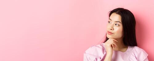 Close-up portrait of romantic asian girl looking aside at logo, thinking or daydreaming, standing against pink background photo