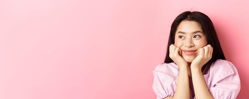 Close-up of excited asian girl listening with interest, smiling amused and looking aside at logo, daydreaming, standing against pink background photo