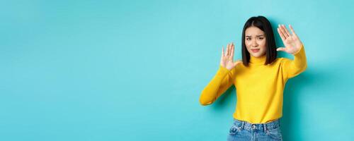 Image of woman feeling uncomfortable in front of camera, cover face with hands and frowning, begging to stop, standing against blue background photo