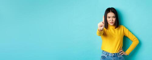 Confident and serious woman tell no, showing extended finger to stop and prohibit something bad, frowning and looking at camera self-assured, standing over blue background photo