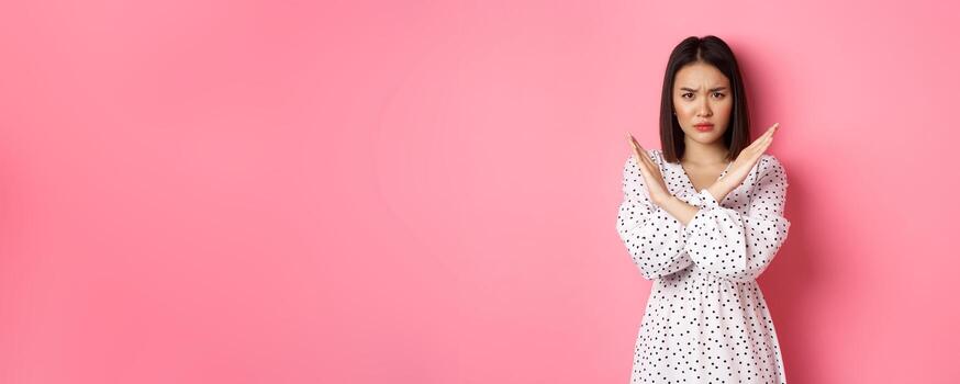 enojado asiático mujer contar detener, haciendo cruzar a prohibir o desaprobar algo, ceñudo disgustado, en pie terminado rosado antecedentes foto