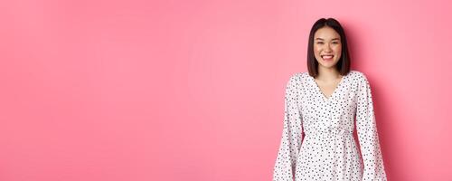 Happy korean woman in dress looking at camera, smiling and laughing with sincere expression, standing over pink background photo