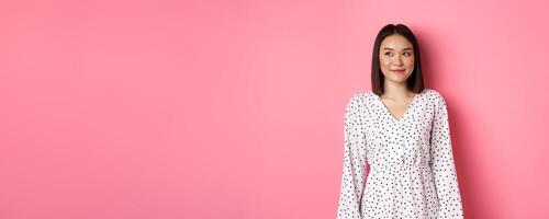 Cute asian woman smiling, looking left at copy space, standing on romantic pink background photo