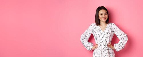 Beautiful korean lady in dress looking at camera and smiling, waiting for something, standing against pink background photo