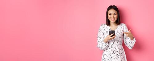 Beautiful young asian woman in romantic dress using smartphone, smiling and pointing at mobile phone, standing over pink background photo