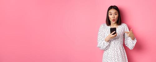 Online shopping and beauty concept. Amazed asian woman pointing finger at mobile phone, recommending smartphone app, standing over pink background photo