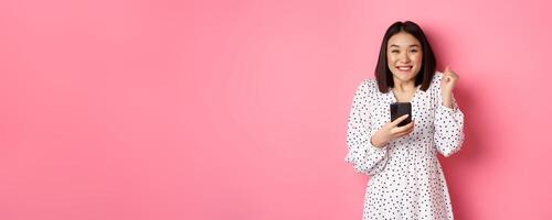 Online shopping and beauty concept. Excited asian woman winning in internet, holding smartphone and rejoicing, smiling happy and celebrating, standing over pink background photo