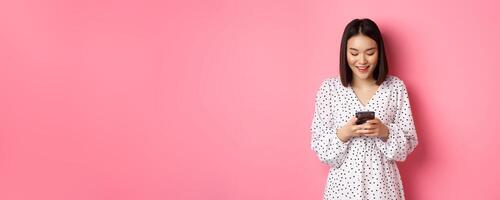 Beautiful asian lady reading message and smiling, using mobile phone, standing in cute dress against pink background photo