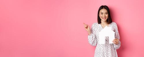 Real estate. Excited asian woman showing paper house model, pointing left at copy space, standing over pink background photo