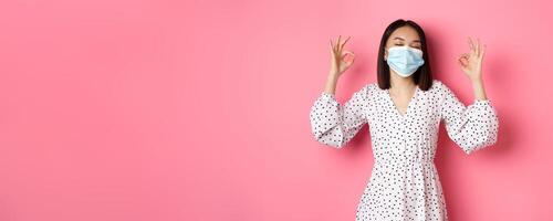 Coronavirus, social distancing and lifestyle concept. Satisfied asian woman in face mask showing okay signs, approve and agree, praising good choice, pink background photo