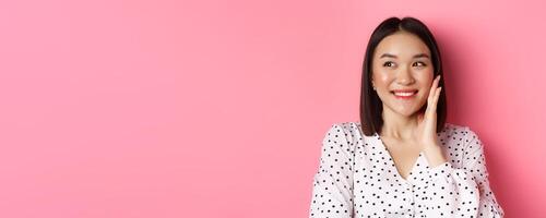 Coquettish asian woman in blushing, touching cheek and looking left amused, standing over pink background photo