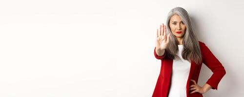 Serious asian woman in red blazer showing stop sign, telling no, frowning and rejecting something bad, standing against white background photo