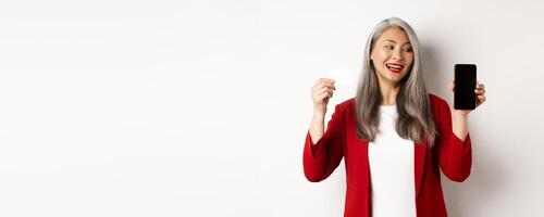 Cheerful asian mature woman showing blank smartphone screen and credit card, concept of e-commerce photo