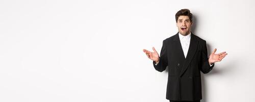 Portrait of confused and worried handsome man in suit, looking at something strange, spread hands sideways and standing puzzled against white background photo