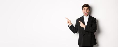 Portrait of excited and happy man in suit showing advertisement, pointing fingers at upper left corner and smiling amazed, standing over white background photo